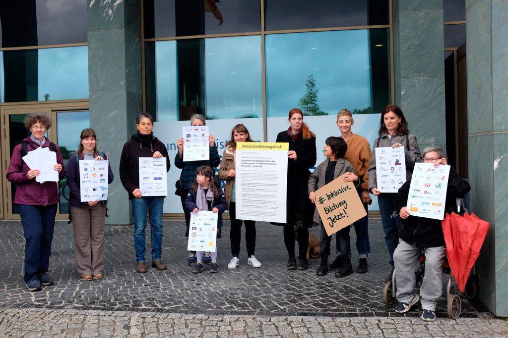 Eine Gruppe von Aktivistinnen  stehen vor dem Bundesministerium für Bildung und Forschung, In Ihren Händen halten sie den Offenen Brief #InklusiveBildungJetzt! und Blätter mit den Logos der 130 unterzeichnenden Verbände und Organisatioen.